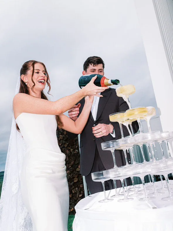 Alex and Will laughing while pouring champagne into a tower of glasses