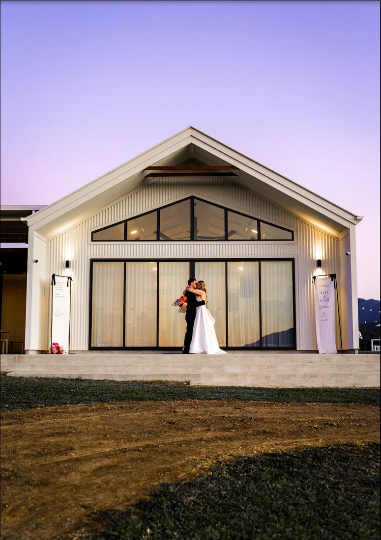 Gabi and Alec kissing on the venue steps at dusk, the reception building lit up behind them