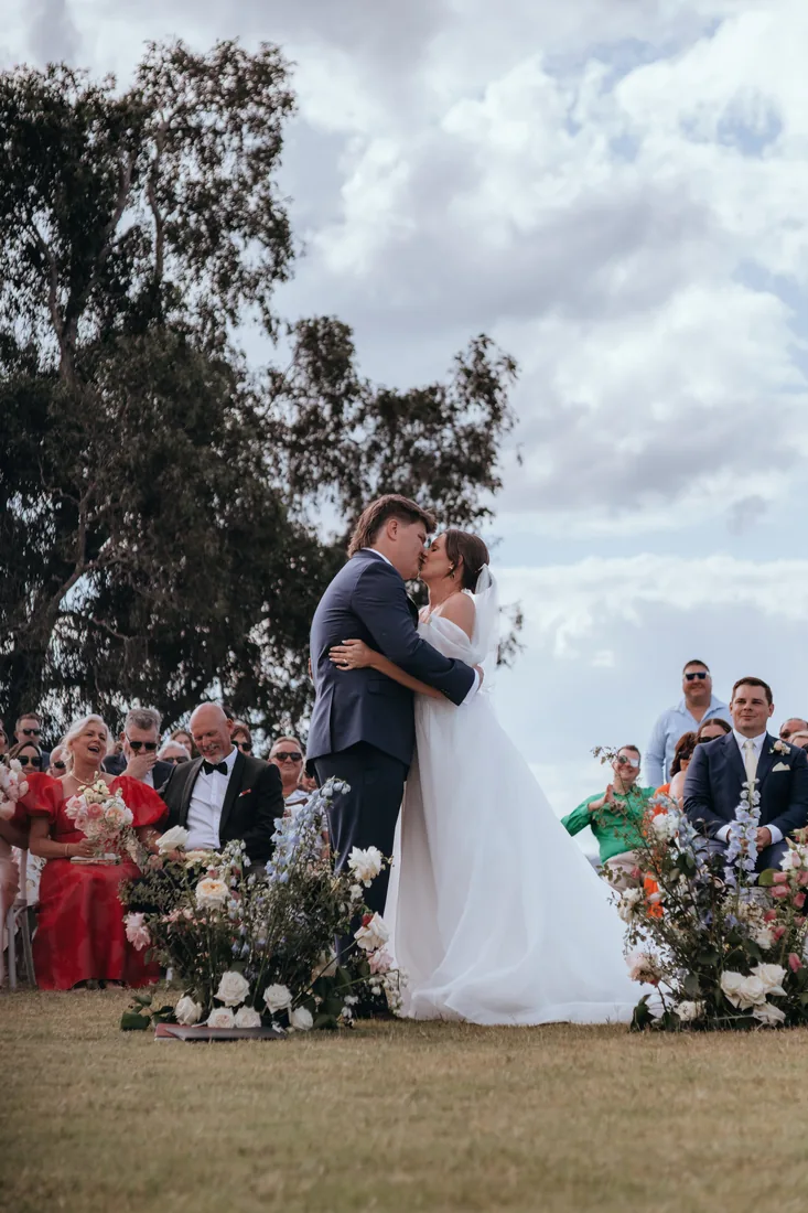 Jorjia and James sharing their first kiss as married couple, guests seated among florals