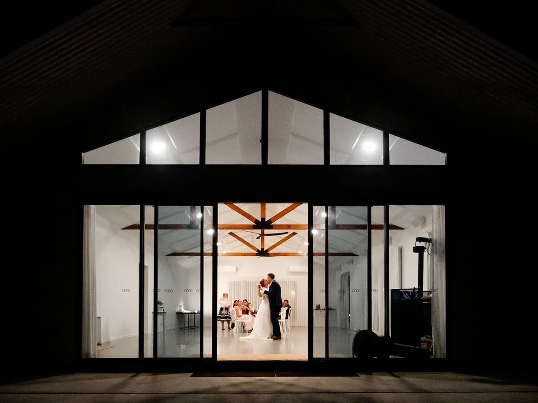 Night photo of the reception venue lit up, Leisa and Russell sharing their first dance visible through the glass doors