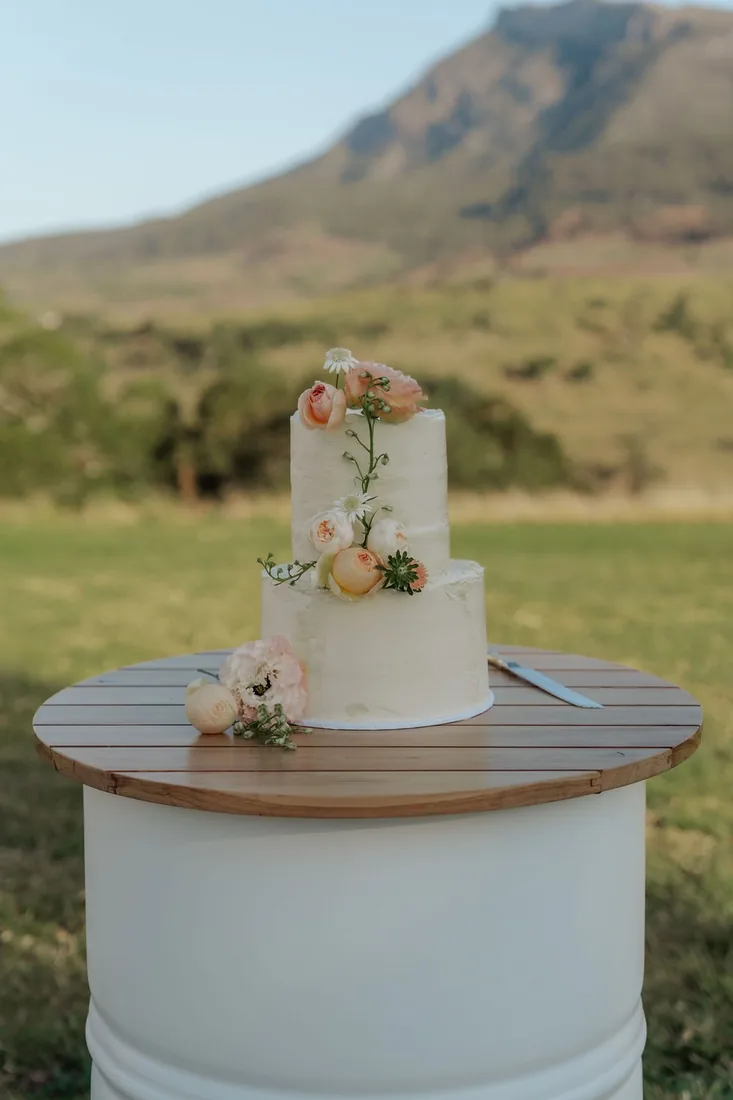 Semi-naked wedding cake with peach and white florals on a timber cake stand, mountains behind