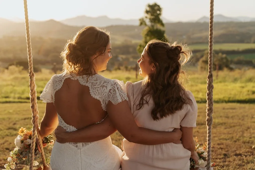 Bride and bridesmaid embracing on a wooden swing at golden hour