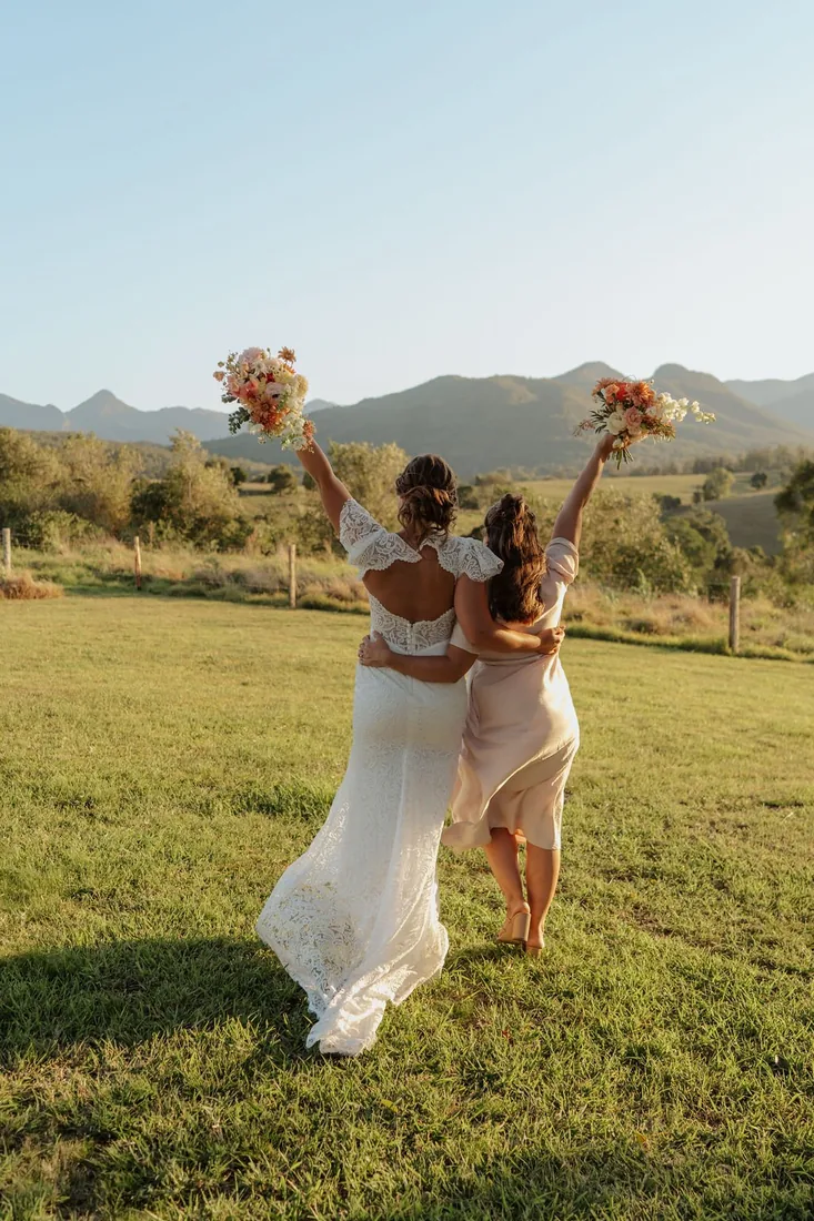 Bride and bridesmaid running across the lawn with bouquets raised in celebration