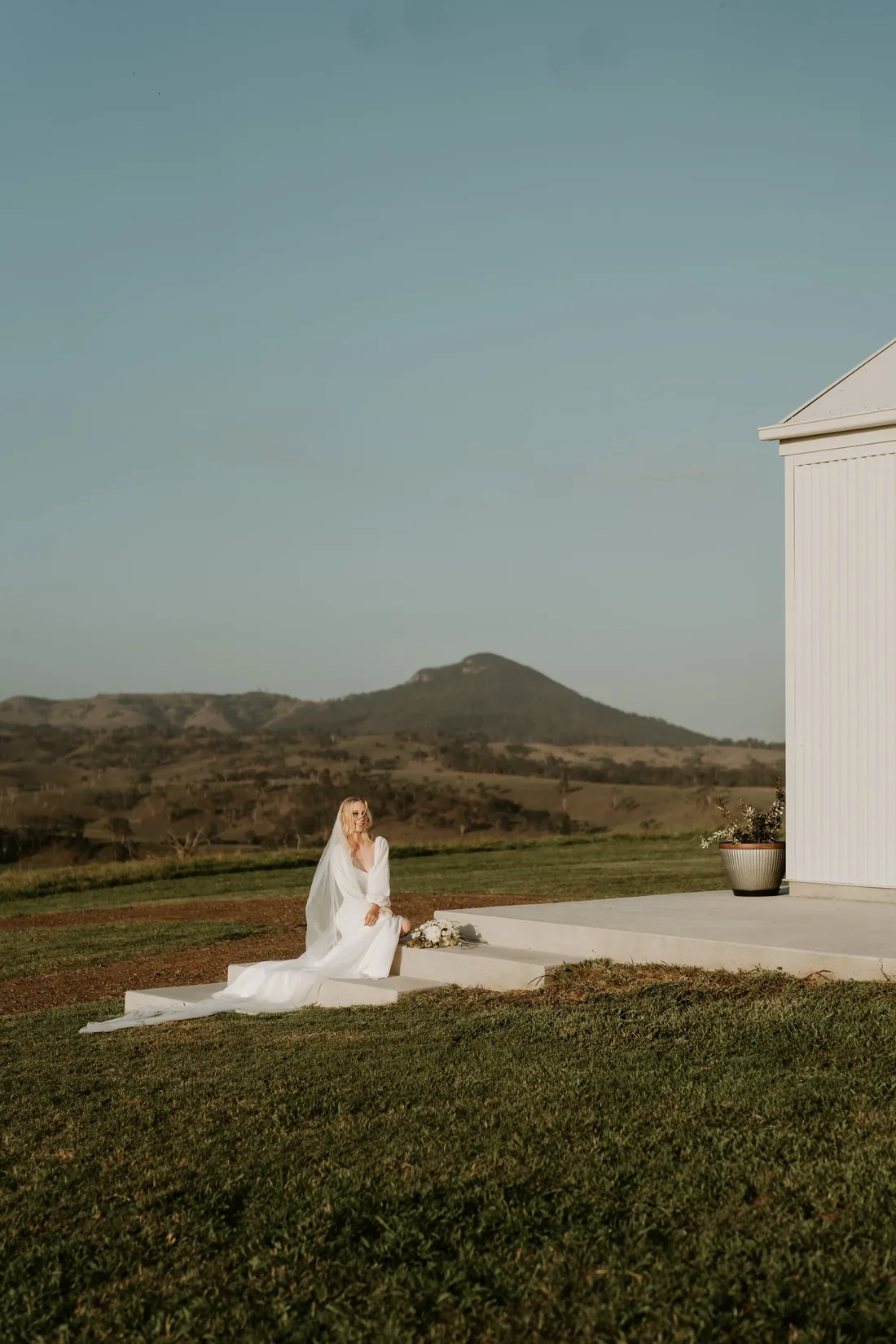 Bride sitting on the venue steps in a long-sleeve gown, mountain views behind her