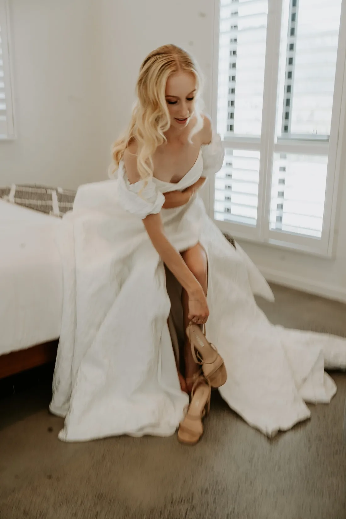 Bride sitting on the edge of a bed slipping into sculptural nude heels