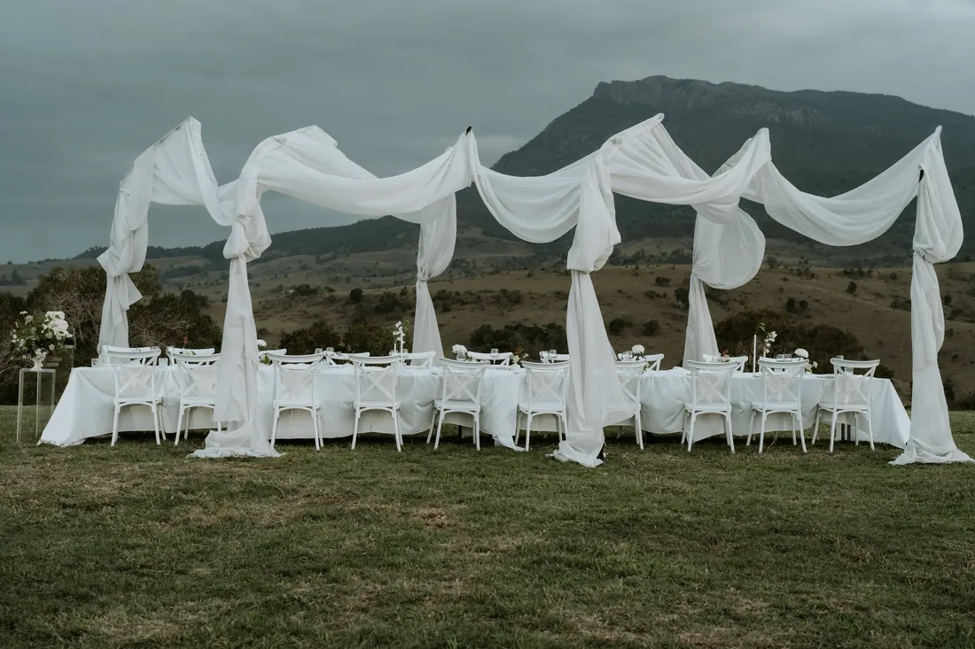 Sheer draped reception arbour and long table set for dinner under an overcast sky, mountain backdrop