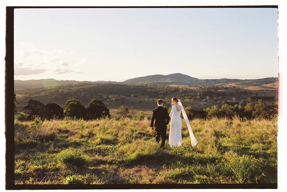 Sally and Joshua walking hand in hand through golden grass at sunset with mountains behind them
