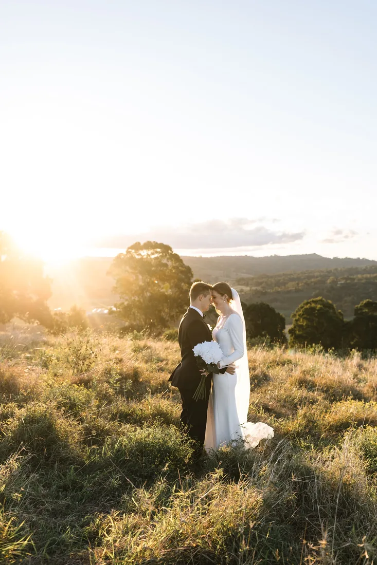 Sally and Joshua embracing at golden hour in tall grass, backlit by the setting sun