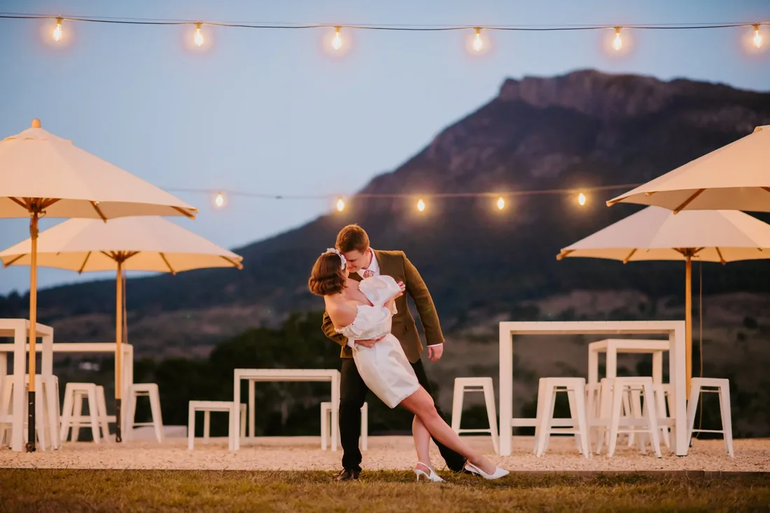 Couple sharing a dip kiss on the lawn at dusk beneath festoon lights at Maroon Hill Estate